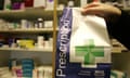 A prescription paper bag held by a hand with various drugs in the background