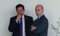 Two men in suits stand and talk in front of a white wall outside