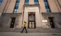 person wearing yellow top and black pants walks past tan building