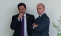 Two men in suits stand and talk in front of a white wall outside