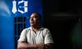 A Mexican man in a T-shirt in front of a blue wall with a barred up window