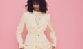 A woman with curly dark hair wearing a pale yellow suit and top, and standing in front of a pink background