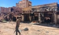 A man wearing camouflage patterned clothes and holding an automatic rifle walks past a burnt-out building.